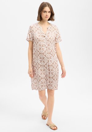 Frau läuft, trägt ein knieumspieltes kurzärmeliges Kleid mit beige-weißem Muster und beigen Pantoletten auf weißem Hintergrund.