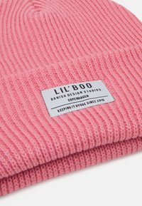 Beanie rosa a coste con risvolto, dotato di un'etichetta rettangolare con testo nero che fornisce informazioni sul marchio e sull'origine.