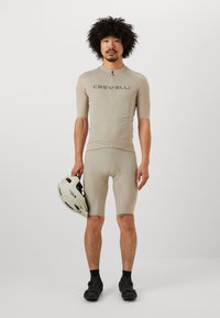 Ensemble de cyclisme beige avec des manches courtes et un zip à l'avant, arborant le logo "Castelli", associé à un short assorti et un casque léger.