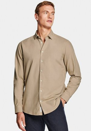 Man draagt beige overhemd met lange mouwen en kraag, staat met één hand in de zak en kijkt naar de zijkant.