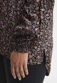 Melna blūze ar ziedu rakstu, garām piedurknēm, ar smocka aprocēm un sānu šķērslinijām; tai ir purpursarkani un dzelteni ziedu akcenti uz teksturētas auduma.