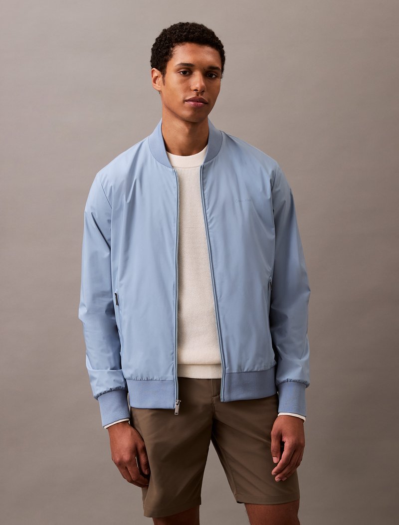 Giacca bomber azzurra in materiale liscio, con chiusura a zip, polsini a costine e un design semplice, indossata sopra un maglione crema.