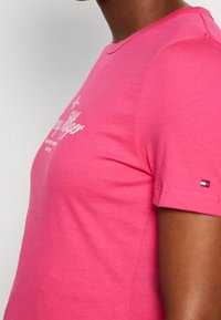 Rosa t-shirt med korte ærmer lavet af bomuld, med hvid tekstdesign og et lille tri-farvet logo på ærmet. Glat tekstur.
