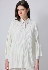 Suéter blanco de punto oversized con un profundo escote en V, mangas largas y textura de canalé vertical. Superpuesto sobre una camisa con cuello.