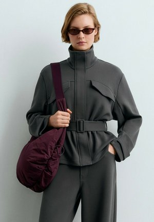 Personne portant une veste ceinturée gris foncé et un pantalon, portant un grand sac à épaule violet, et portant de petites lunettes de soleil foncées sur un fond uni.