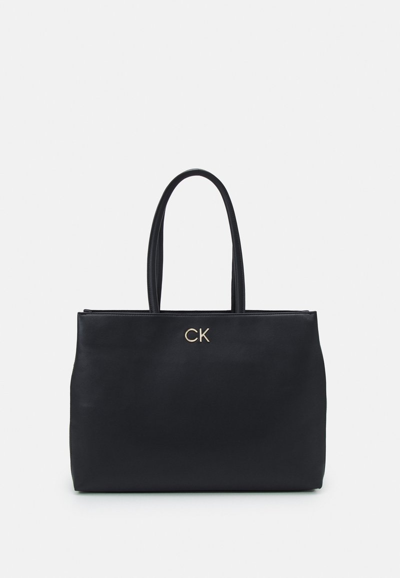 Czarna skórzana torba typu tote z dwoma długimi uchwytami, ozdobiona złotym logo „CK” na przodzie. Gładka tekstura i usztywniony kształt.
