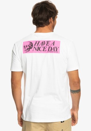 Homme aux cheveux foncés et tatouage au bras porte un t-shirt blanc avec un rectangle rose, une tête de mort noire et le texte « HAVE A NICE DAY » dans le dos.
