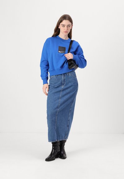 Sweat à capuche bleu avec une poche patch noire, associé à une jupe en denim longue taille haute. Le mannequin porte des bottines noires et tient un sac à main noir.