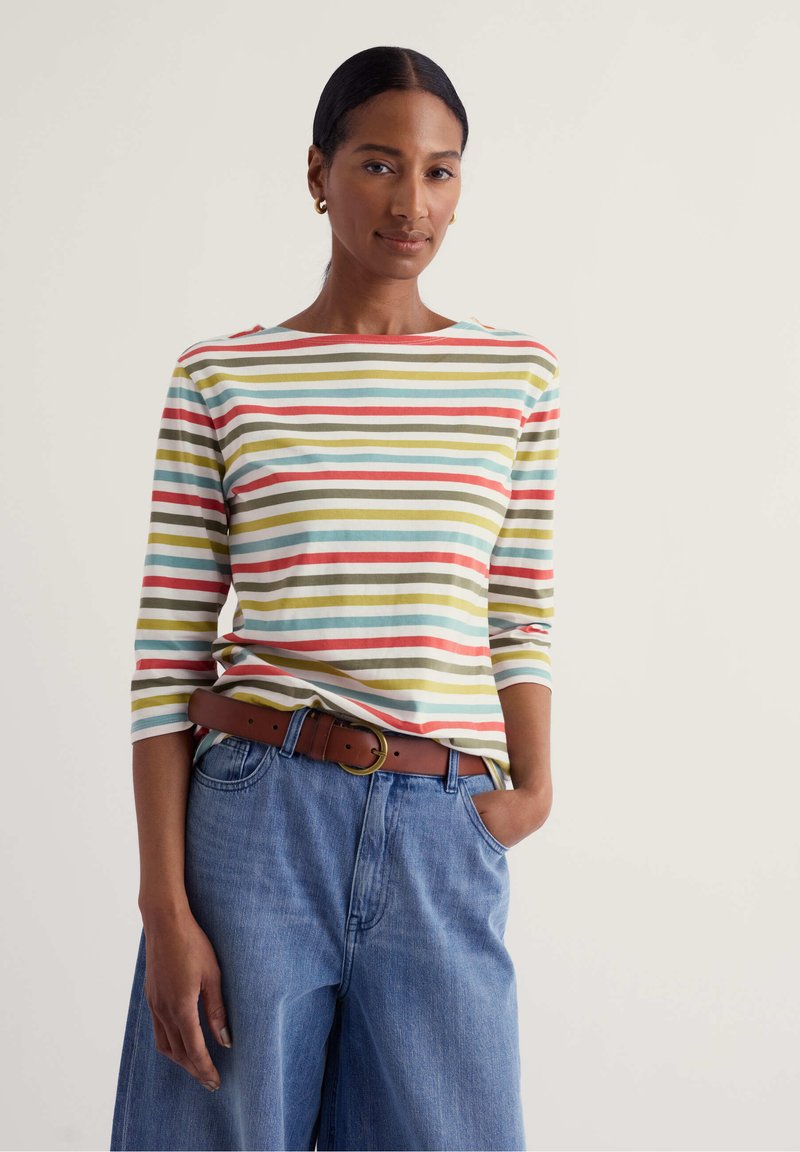 Vrouw met donker haar in een staart draagt een gestreept shirt met lange mouwen, blauwe wijde jeans en een bruine riem, staand met één hand in de zak.