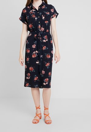 Kvinde iført knælængt sort blomsterkjole med knapper og taljebånd, kombineret med orange sandaler med stropper og blokhæl.