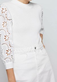 Kvinna iklädd en vit stickad ärmlös topp med ögla blommönster och vita kjol med flätad midja, hand i fickan.