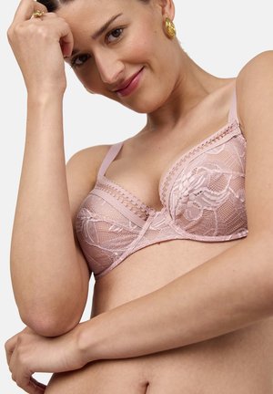 Donna sorridente che indossa un reggiseno di pizzo rosa chiaro, appoggiando la testa sulla mano sinistra, con orecchini a cerchio dorati e un anello d'oro.