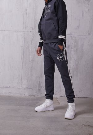 Personne portant un sweat à capuche noir Nike et un pantalon de jogging assorti avec des bandes blanches, debout sur un sol en béton devant un mur gris texturé.