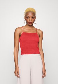 Top cropped vermelho com franzido e alças finas, apresentando um design texturizado e pregueado, com bainha em forma de ondas. Combinado com calças de cintura alta de cor clara.