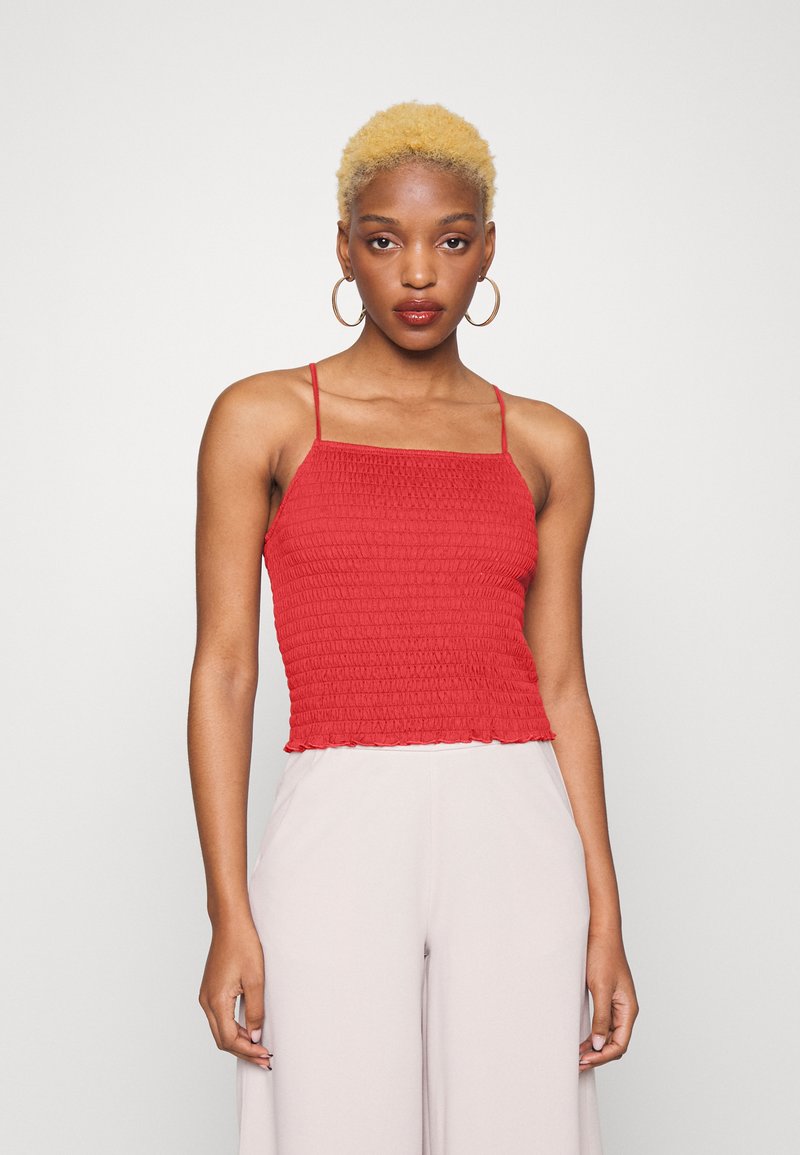 Top cropped vermelho com franzido e alças finas, apresentando um design texturizado e pregueado, com bainha em forma de ondas. Combinado com calças de cintura alta de cor clara.
