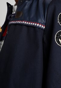 Marinblå jacka med läderaxelpanel, röd, vit och blå vävd trim, broderade cirkulära lappar och marinblå dragsko vid kragen.