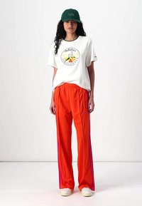 Balta marškinėliai su spalvingu Adidas logotipu, derinami su oranžinėmis sportinėmis kelnėmis su rožinėmis šoninėmis juostelėmis ir baltais sportbačiais. Žalias kepurė.