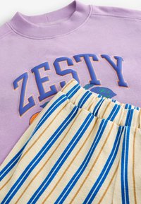 Fialová mikina s nápisem "ZESTY" a grafikou; spárována s pruhovanými béžovými šortkami s modrými a hnědými pruhy a texturovanou tkaninou.