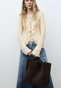 Licht beige gerimpelde blouse met lange mouwen, gecombineerd met blauwe denim. Houdt een donkerbruin suède schoudertas vast. Gouden sieraden als accenten.