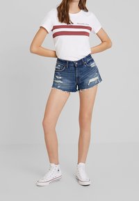 Niebieskie dżinsowe szorty z postrzępionymi detalami i wystrzępionym dołem, zestawione z białym t-shirtem z bordowymi paskami. Noszone z białymi wysokimi trampkami.