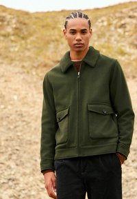 Young man with braided hair wearing a dark green zip-up jacket with large buttoned pockets standing outdoors on rocky ground.
