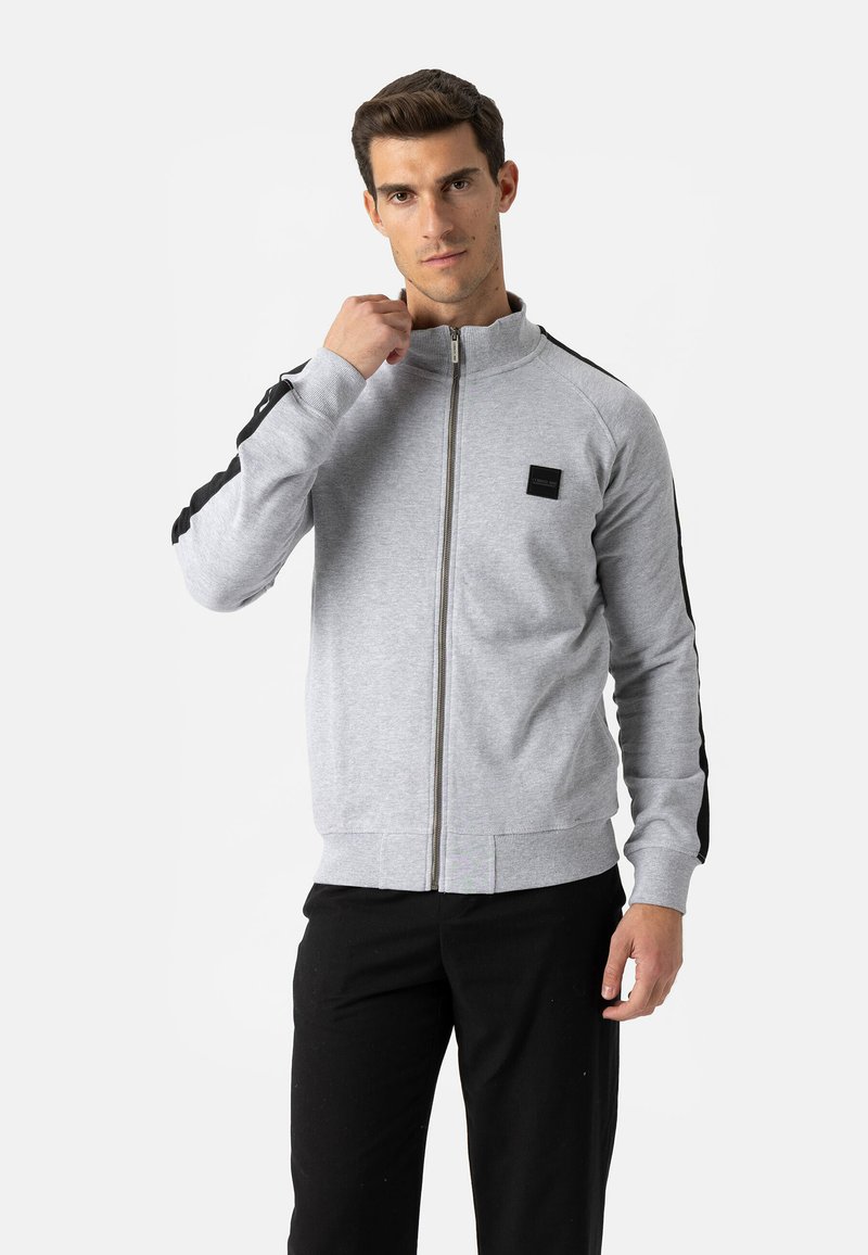 Grijze sweatvest met rits en hoge kraag, zwarte zijstrepen en een klein logoplaatje op de borst. Gladde stof en nauwsluitend ontwerp.