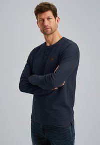 Mornarsko plava majica dugih rukava henley od teksturiranog materijala, s gumbima na prednjem dijelu i malom oznakom brenda na prsima.