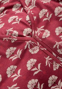 Roter Stoff mit einem Blumenmuster, das weiße Blumen und grüne Akzente zeigt. Das Design umfasst Blätter und ist strukturiert mit sichtbaren Nähten.