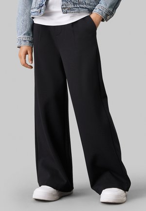Pantaloni neri a gamba larga realizzati in tessuto morbido con una texture liscia, caratterizzati da un dettaglio di cucitura anteriore e abbinati a scarpe da ginnastica bianche.