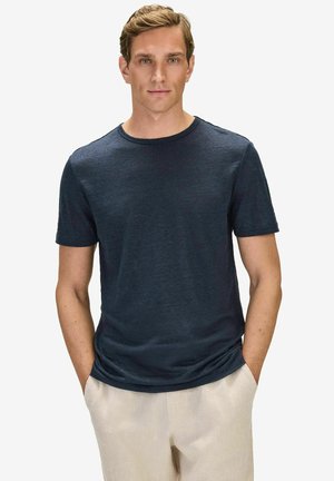 Man wearing a plain dark navy short-sleeve t-shirt and light beige pants, standing with hands in pockets against a white background.