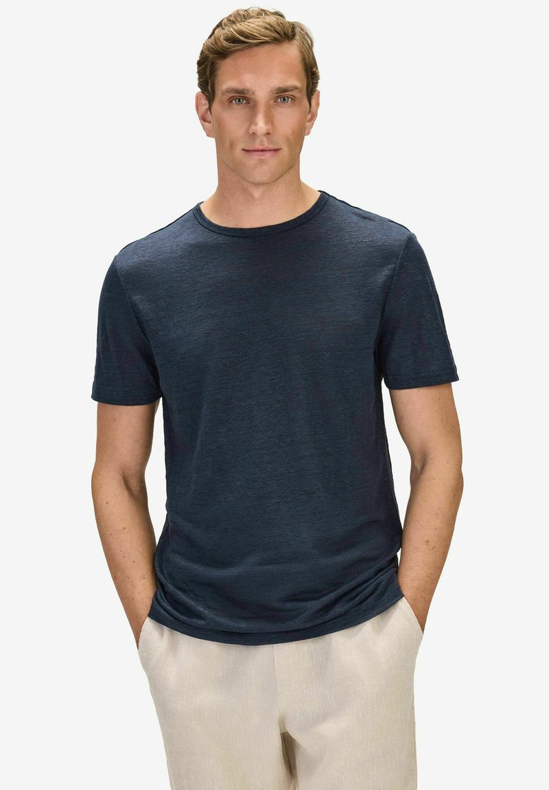 Homme portant un t-shirt à manches courtes bleu marine uni et un pantalon beige clair, debout les mains dans les poches sur fond blanc.