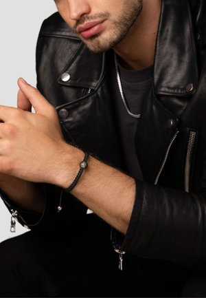 Mand i sort læderjakke iført et sort flettet armbånd med en lille sølvperle på håndleddet og en sølvkæde halskæde.