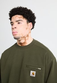Sudadera verde oliva con cuello redondo, que cuenta con un bolsillo en el pecho con cremallera y un parche con el logo de Carhartt. Textura suave, ajuste casual.