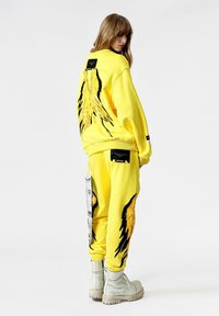 Sudadera amarilla y pantalones con diseños gráficos de alas en negro y amarillo y bolsillos laterales. El conjunto se complementa con botas blancas.