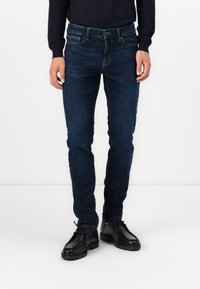 Man som bär mörkblå slimfit-jeans och svarta läderskor med snörning, stående mot en vit bakgrund.