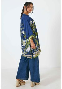 Blauwe bloemen tuniek met groene accenten, voorzien van 3/4 mouwen en een losse pasvorm, gecombineerd met wijde jeans en sandalen met hak.