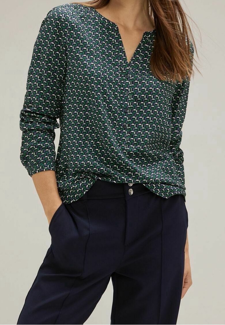 Femme portant un chemisier à manches longues vert foncé à motifs, rentré dans un pantalon bleu foncé taille haute, les mains dans les poches.