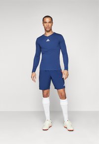 Maglietta sportiva a maniche lunghe color navy con logo Adidas, abbinata a pantaloni corti coordinati. Calzini bianchi e scarpe leggere completano l'outfit sportivo.