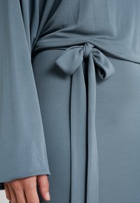 Jasnoniebieska sukienka z miękkiej, fakturowanej tkaniny, z wiązanym detalem w pasie i długimi, zwiewnymi rękawami.