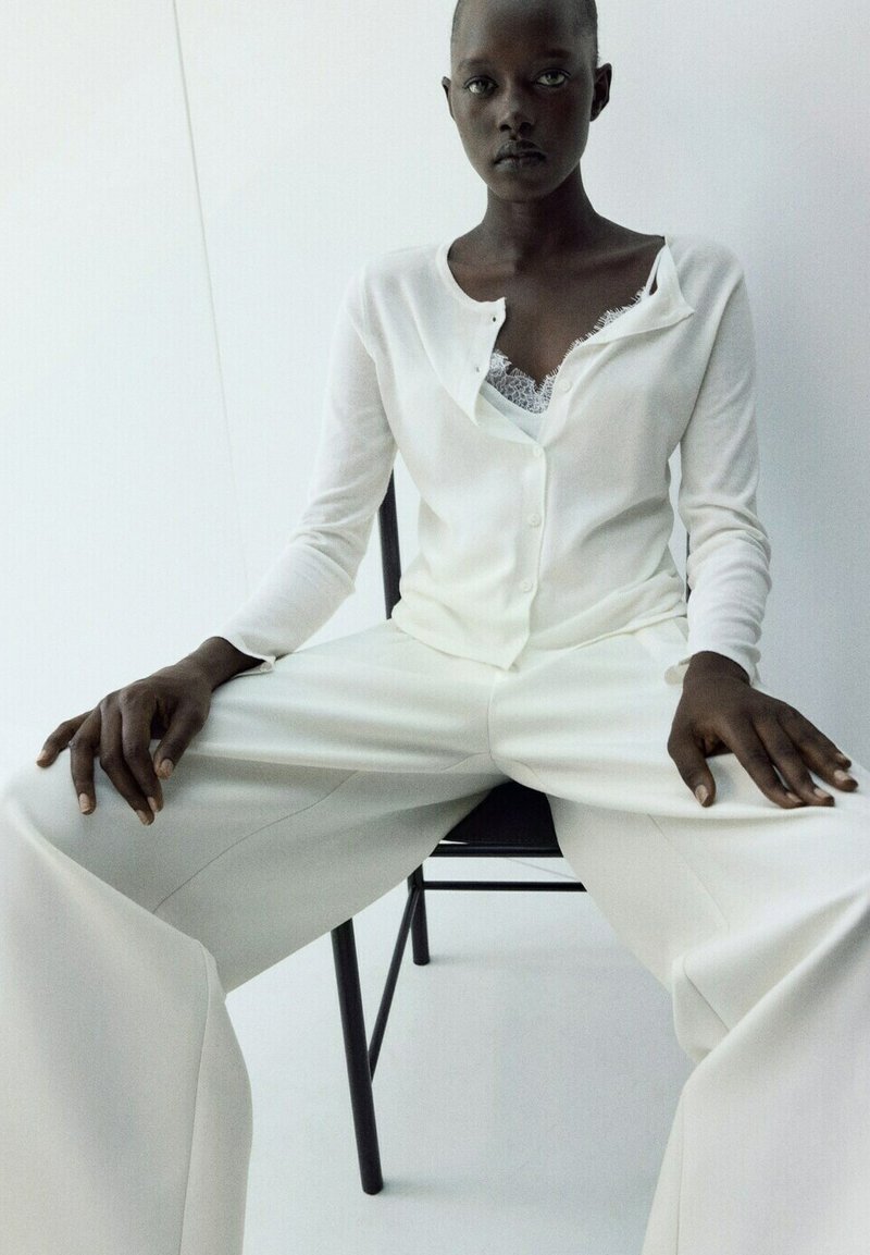 Personne à la peau foncée assise sur une chaise noire, portant un pantalon blanc ample, un cardigan blanc boutonné avec un top orné de dentelle en dessous, faisant face vers l'avant.