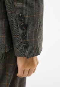 Pilkas languotas švarkas su rankovėmis, ant kurių yra keturi juodi mygtukai ir oranžinės akcentinės linijos, einančios per medžiagos tekstūrą.