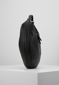 Czarna torebka na ramię z teksturowanej skóry, z górnym paskiem i zamkiem, stojąca pionowo na białym piedestale na szarym tle.