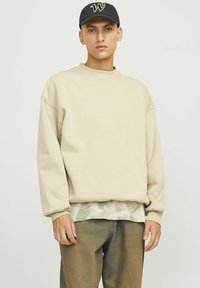 Sudadera beige con hombros caídos y mangas largas, sobre una camisa ligera de estampado de camuflaje, combinada con jeans marrón claro. Gorro negro.