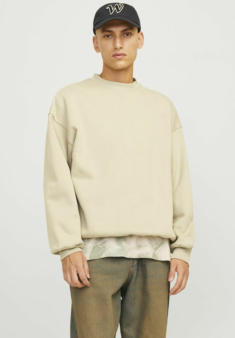 Beige sweatshirt med sänkta axlar och långa ärmar, lager-på-lager över en ljus camoshirt, matchad med ljusbruna jeans. Svart keps.