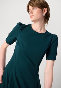 Teal fitted dress with short puff sleeves; features gold accents on cuffs and a smooth, textured fabric. Round neck and flowing silhouette.
