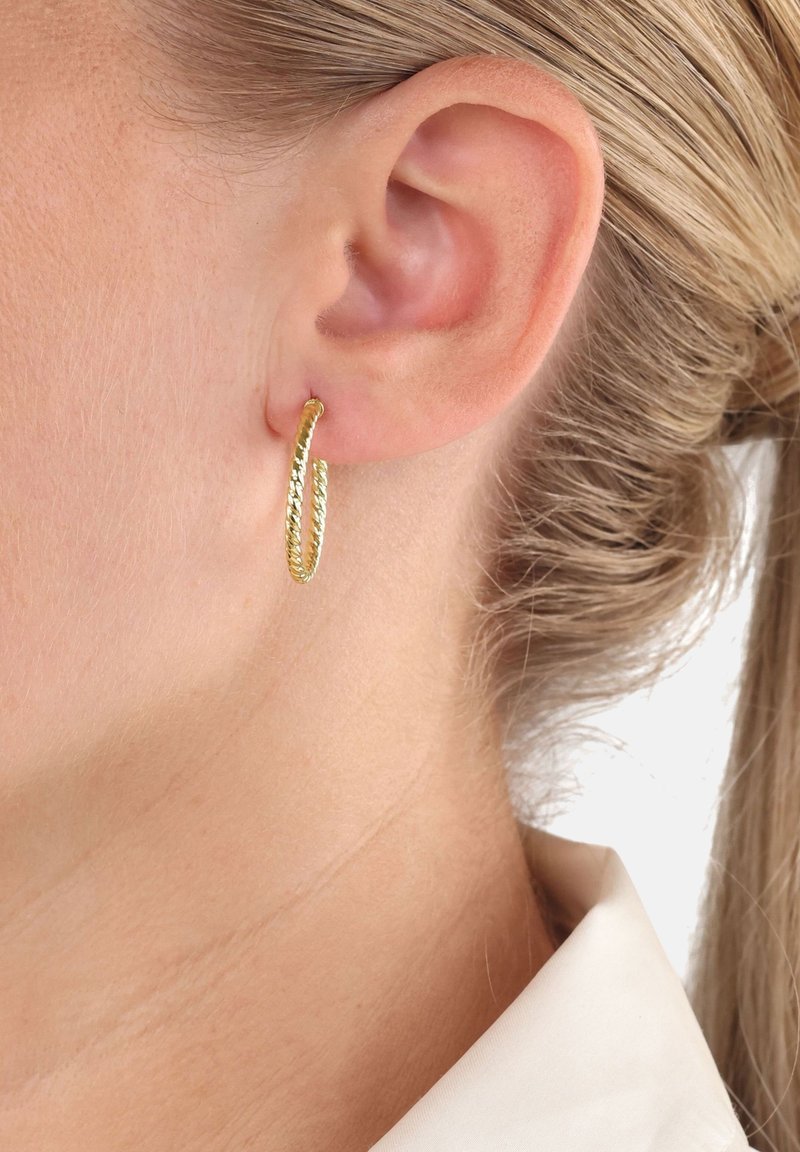 Pendiente de aro con textura dorada y diseño retorcido colgando de la oreja, mostrando un acabado brillante y forma abierta.
