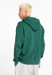 Felpa verde con cappuccio realizzata in tessuto morbido. Presenta una vestibilità comoda, polsini e orlo a coste. Indossata con pantaloni della tuta grigi.