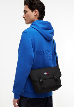 Noor mees sinises kapuutsiga sviitri ja tikitud tekstiga, kandes musta Tommy Jeans'i logoga risti üle keha kotti, vaadates vasakule valge tausta ees.