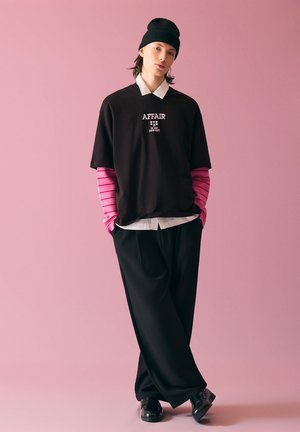 Jeune personne portant un bonnet noir, un t-shirt noir « AFFAIR » sur un sous-pull rayé rose à manches longues, col blanc, pantalons larges noirs, debout devant un fond rose.