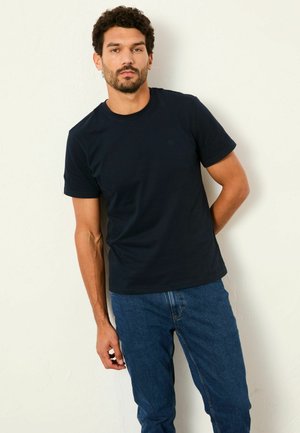 Muž s kučeravými vlasmi a bradou, oblečený v jednoduchom čiernom tričku a modrých džínsoch, stojaci opretý o bielu textúrovanú stenu.